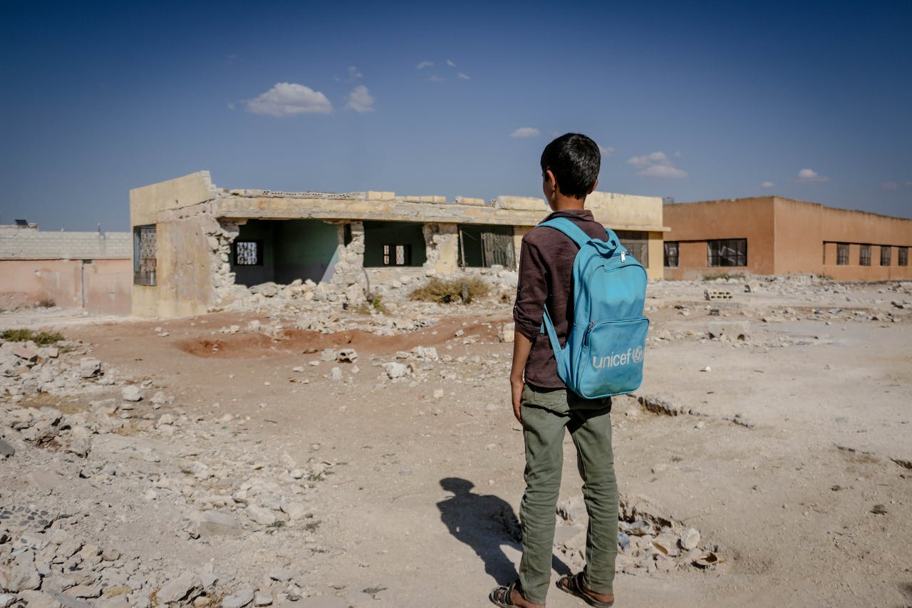 A child with a backpack stands in front of a destroyed building under a clear sky.