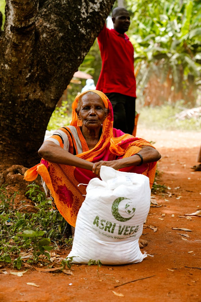 Elderly woman sits by tree with a relief bag labeled 'ASRI VEER'. Rural outdoor setting.