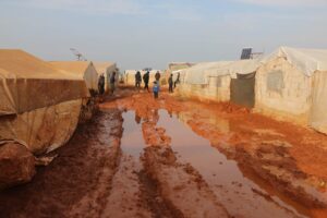 Group of distant adults and kids strolling on wet dirty ground with puddles near worn out tents in poor area