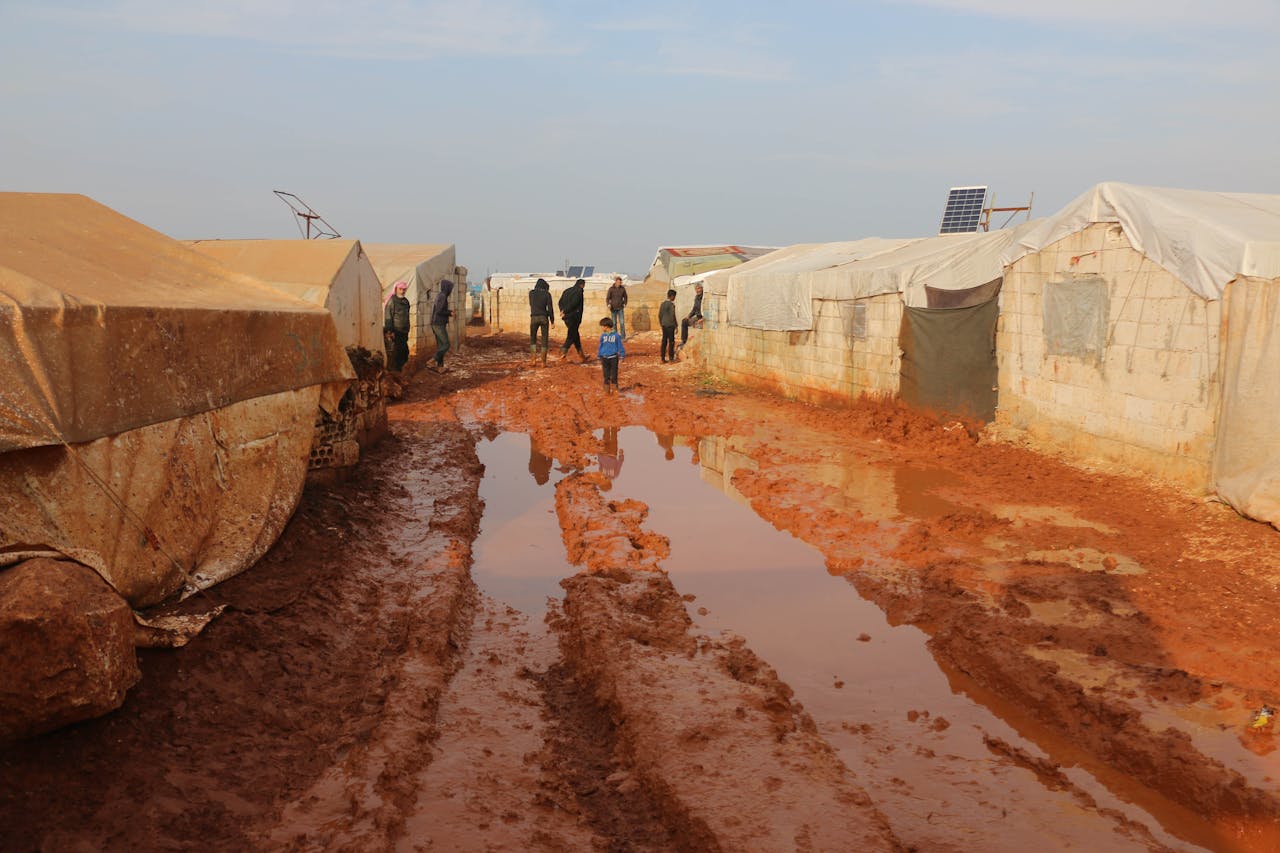 Group of distant adults and kids strolling on wet dirty ground with puddles near worn out tents in poor area